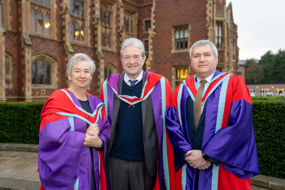 Two male graduates and one female graduate standing together outside a Queen's University Belfast building, smiling and celebrating their accomplishments.